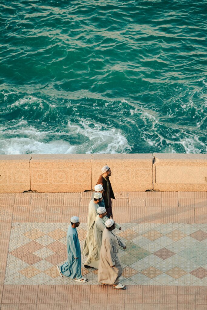 Aerial view of people walking along the Corniche by the sea in Oman, displaying traditional attire.