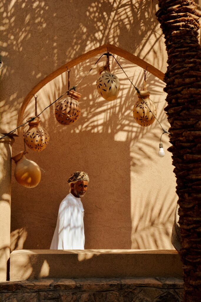 A man in traditional attire stands near an arched doorway in Nizwa, Oman, casting shadows.